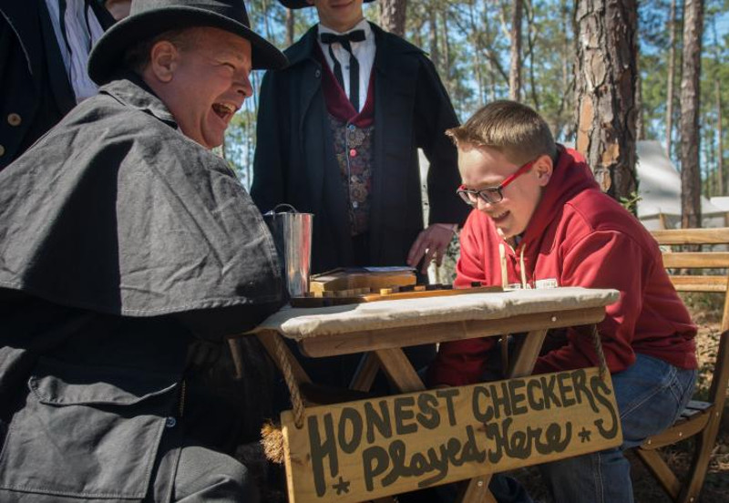 a reenactor and a boy laugh next to a sign reading "honest checkers played here"