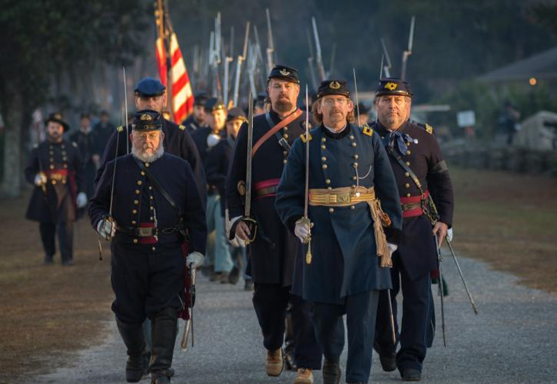 a large group of reenactors dressed as union soldiers march in formation with guns and sabers