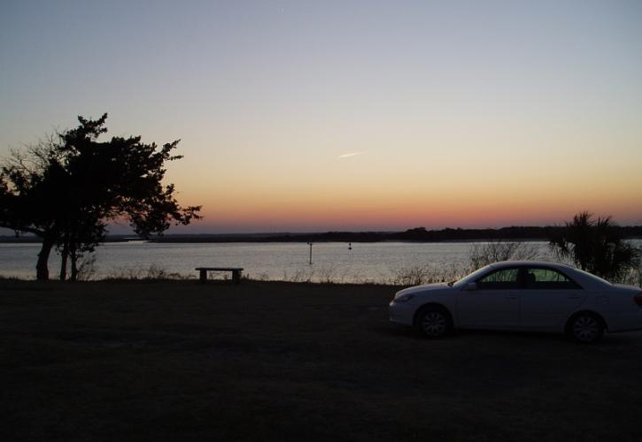 a white car is parked at a grassy plaza with the Amelia River and setting sun in the background