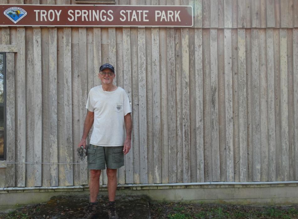 a man in work clothes stands against a wooden fence, below a sign reading "Troy Springs State Park"