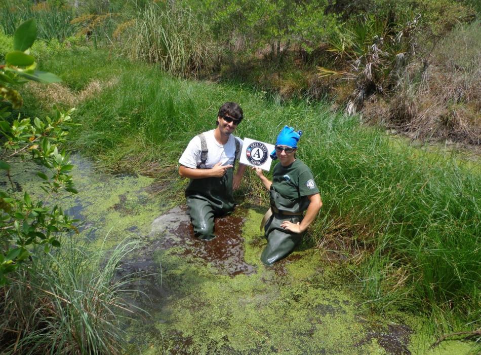 Emily Price and a coworker hold up an AmeriCorps sign while standing knee-deep in water.