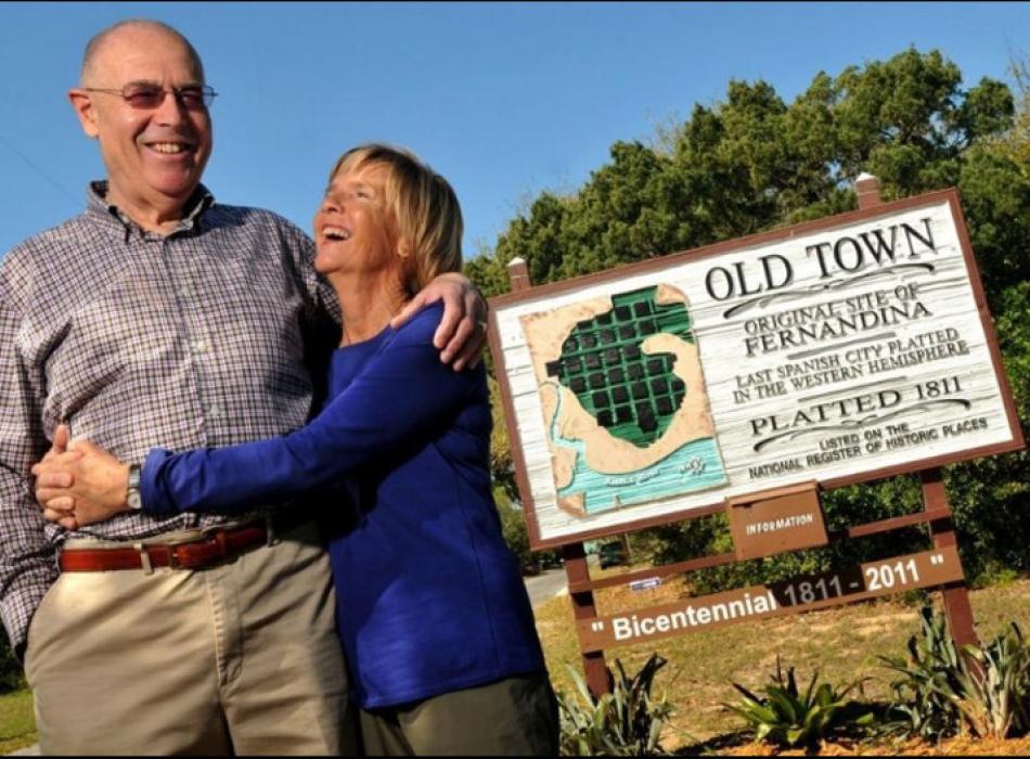 a man and woman hug each other and smile next to a sign for "Old Town Fernandina's Bicentennial"