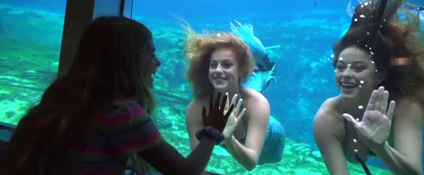 Two mermaids swim while a woman watches from inside the theater.