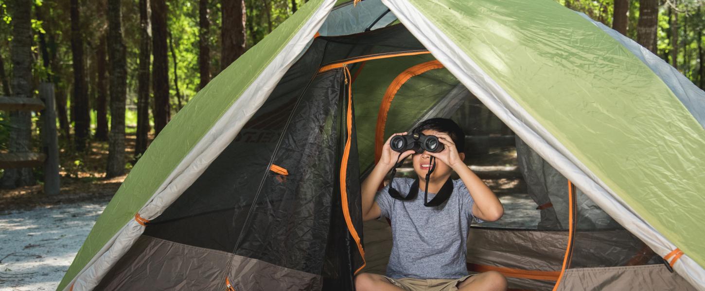 Young boy peers through binoculars from the inside of a tent. 