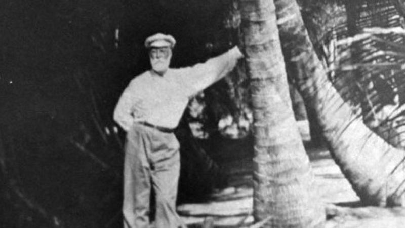 A black and white photo of a man standing next to a large tree.