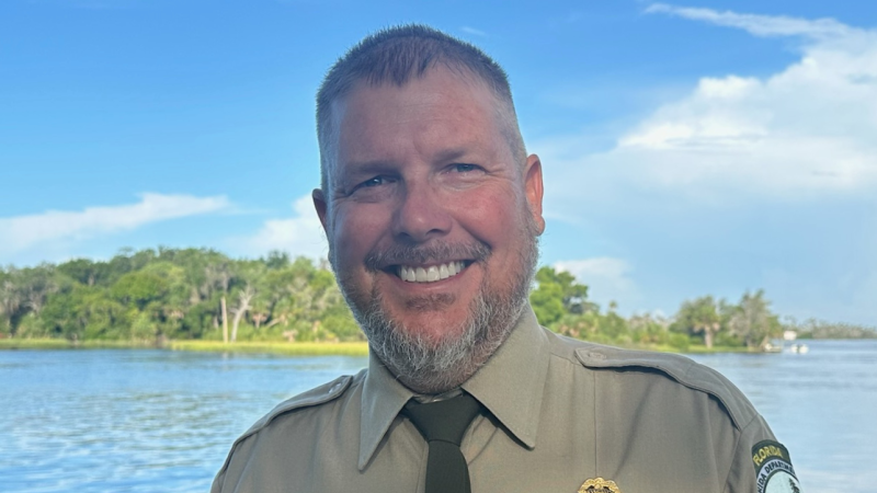 A smiling park ranger wearing class A uniform in front of a lake with trees on the far shore of the lake