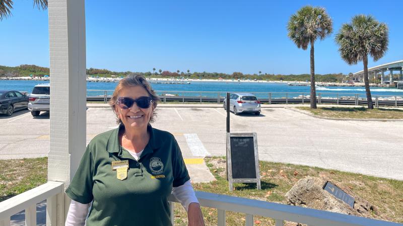 A woman wearing sunglasses, leaning on a railing in front of a parking lot and an inlet.