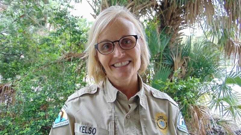 a woman in park service uniform and glasses smiles at the camera in front of a palm tree.
