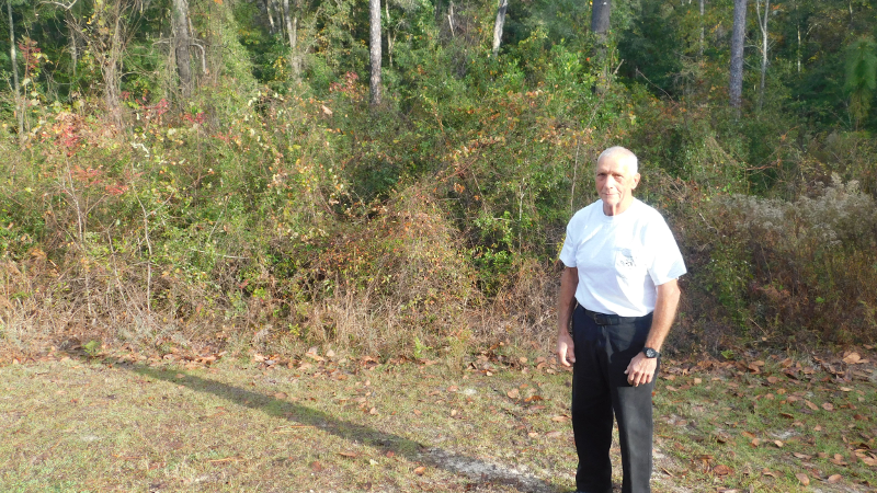 Volunteer Bill Cade stands beside as park trail, with hammock trees in the background.
