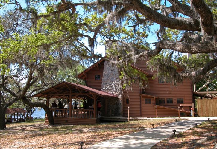 A group of people are seen on deck of lodge building which is surrounded by oak trees draped with spanish moss. 