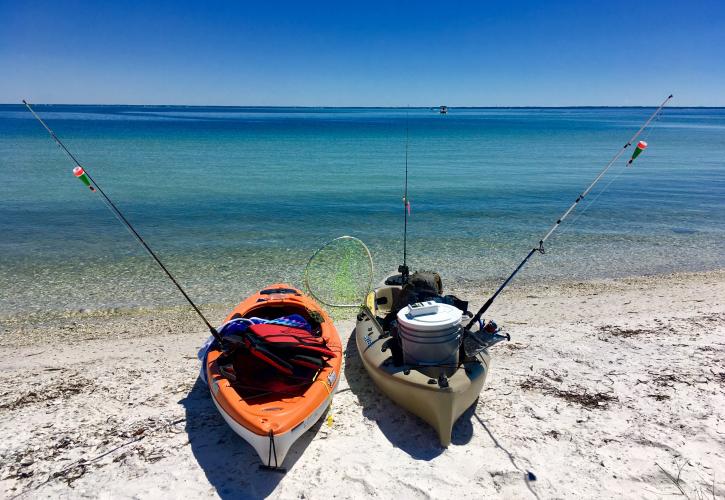 Two kayaks equipped with fishing poles sit on the white sandy beach at the edge of emerald green water.