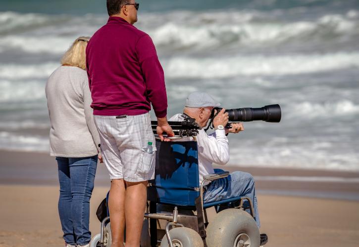 3 visitors on the beach and looking at water, older gentleman is in an all-terrain beach wheelchair taking a photograph