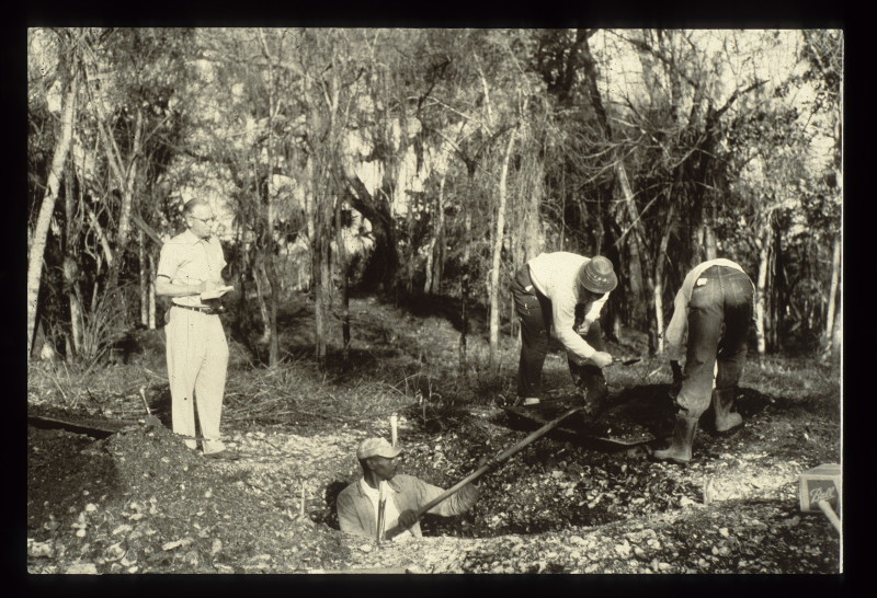 an antique photo of four men digging in the ground and taking notes.