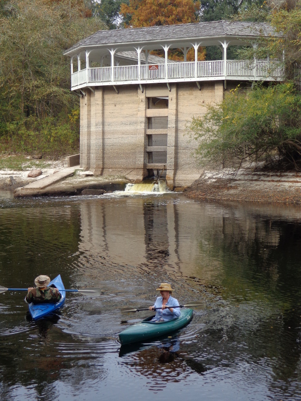 a white stone building with white trim stands in the water as two kayakers paddle by
