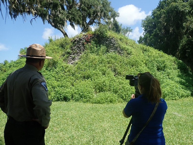 a park ranger and a woman stand in front of a large hill, taking pictures.