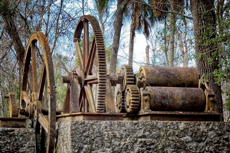 Two rusted metal wheels sit next to three large rusted metal rollers on a machine.