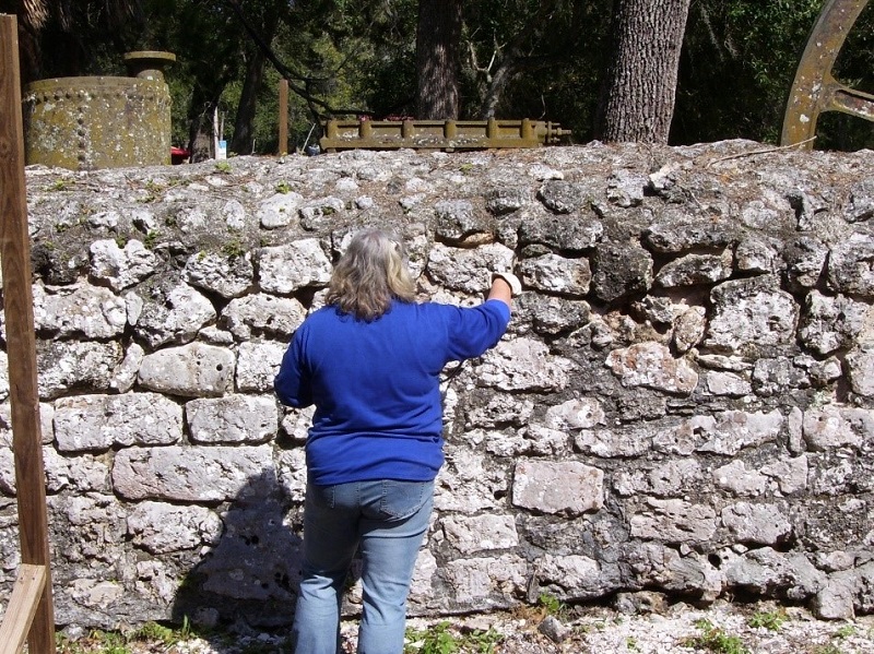A woman in a blue shirt stands painting an old stone wall