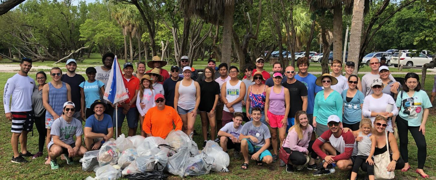 Group photo of dozens of cleanup volunteers at Bill Baggs Cape Florida State Park with trash collected in front. 