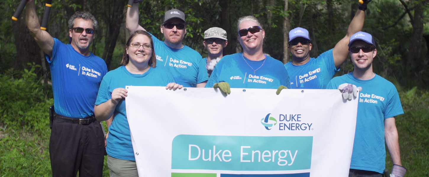 Group of Duke Energy volunteers stand together holding Duke logo sign after a volunteer day. 