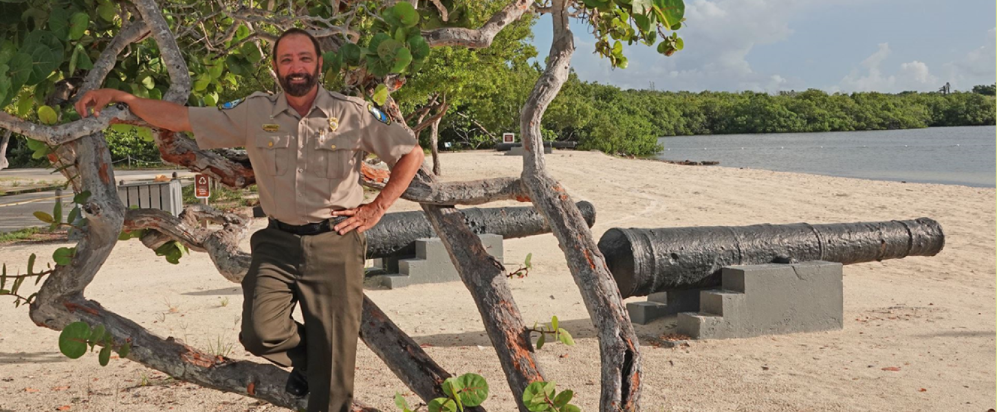 bearded man in Florida Park Service uniform, with background of beach sand, water, green mangroves, and partly cloudy blue skies