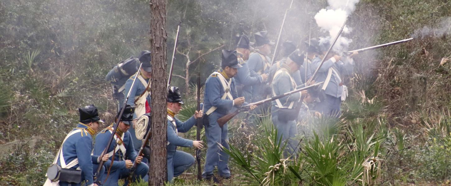 Gun smoke hangs in the air as blue-clad reenactors fire their muskets while crouched in the palmettos.