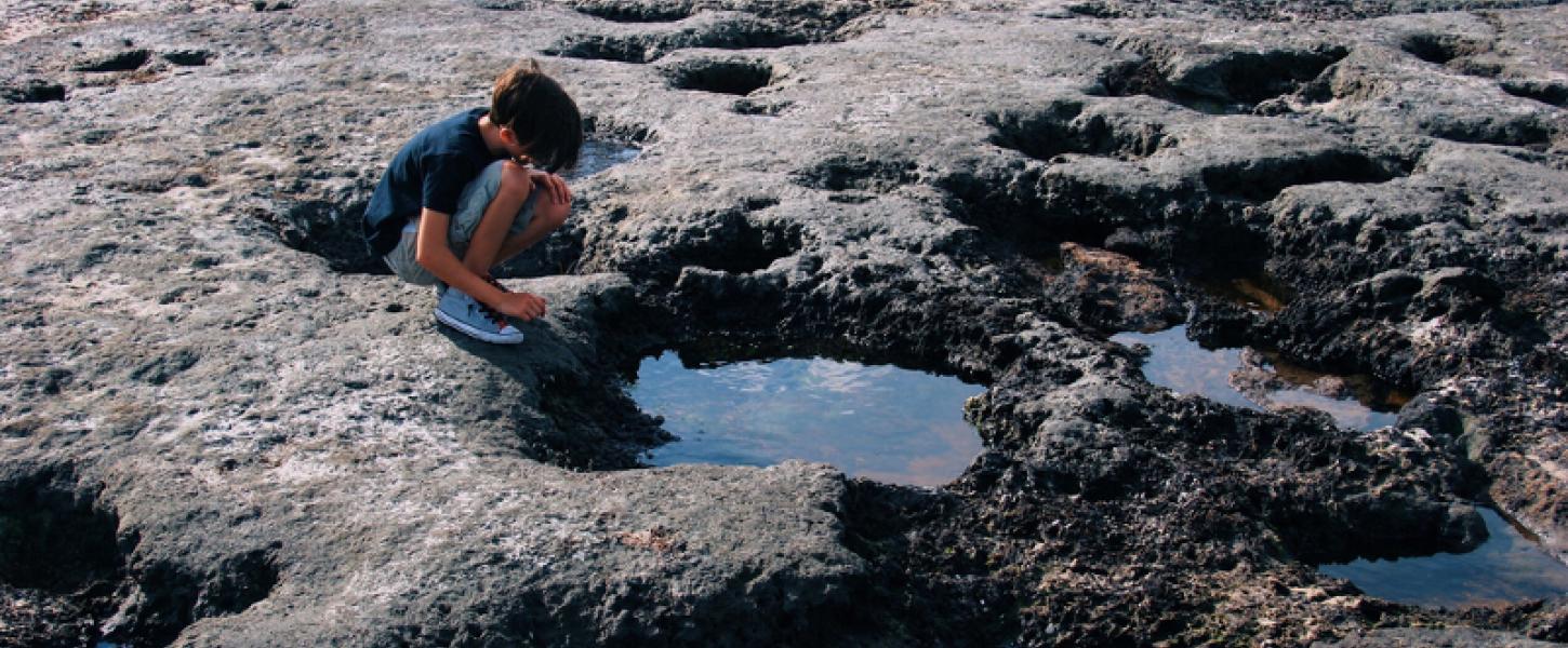 a boy crouches on the edge of a pool formed by hardened black dirt on a beach