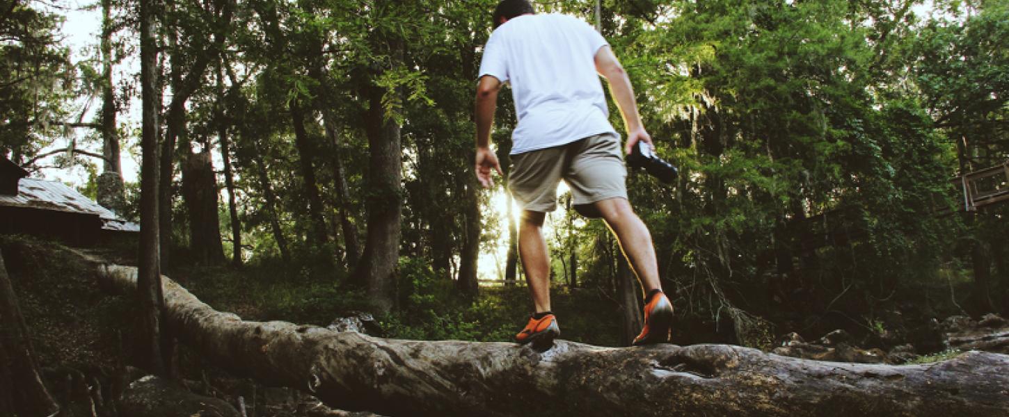 a man in a white shirt and shorts climbs on downed a tree branch carrying a water bottle