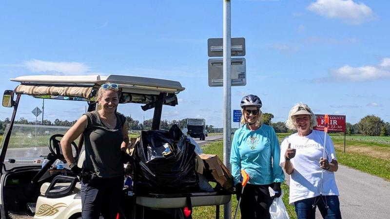 Three women stand on a paved trail next to a sign and a golf cart holding trash bags.