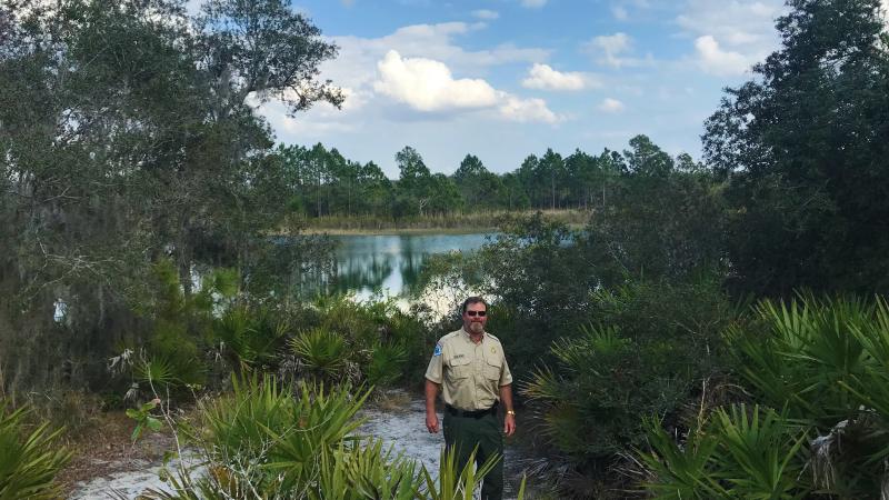Park Manager Andy Noland standing at Catfish Creek surrounded by saw palmetto, oaks, and a lake behind him