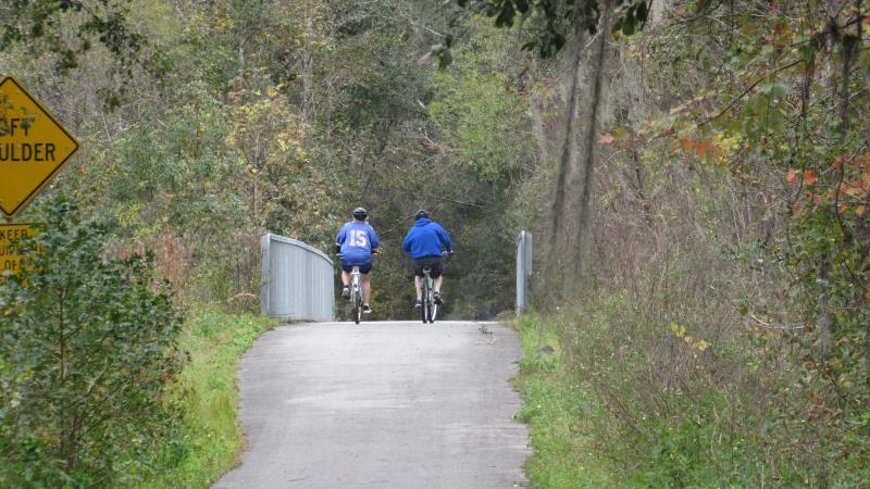 Two people on bicycles ride along a paved trail surrounded by trees. 