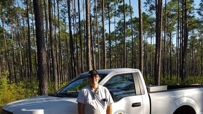 Volunteer Jeff Junior stands in front of park truck and forest of trees. 