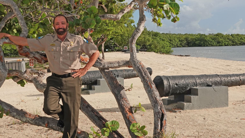 bearded man in Florida Park Service uniform, with background of beach sand, water, green mangroves, and partly cloudy blue skies