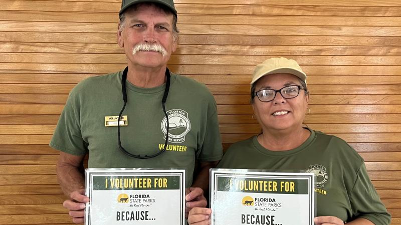 Pam and James hold up signs about why they volunteer: "meeting people" and "sharing barrier free resources."