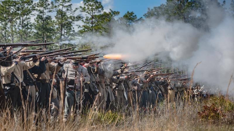 reenactors dressed as confederate soldiers stand in a long line firing guns