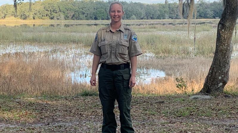 a park ranger stands on a lake shore, surrounded by trees and golden grasses.