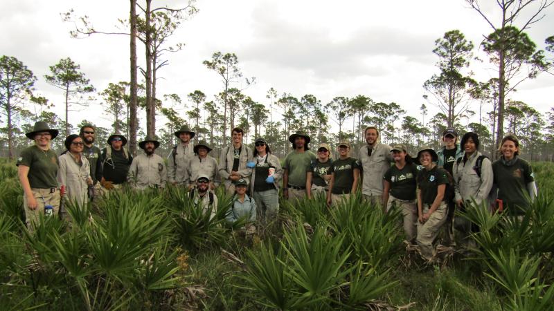 A group of FLCC members pose in a pine scrub landscape, wearing khaki and green uniforms and smiling. 