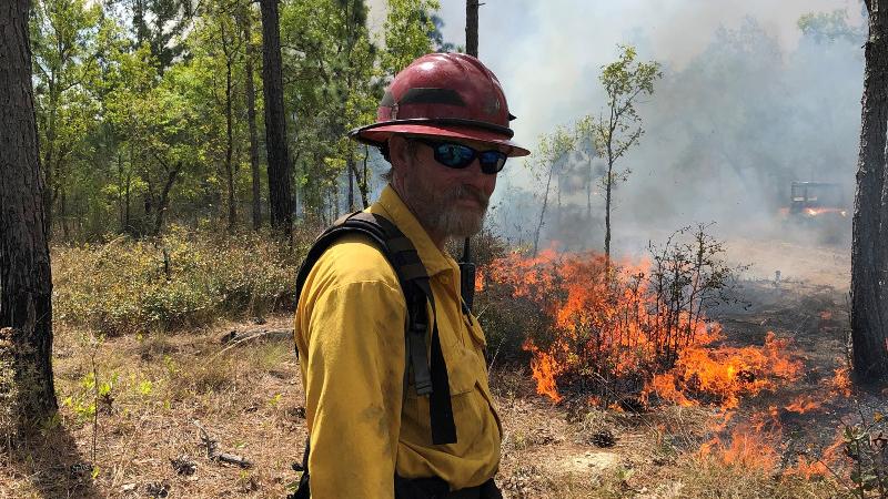 a man in protective gear stand sin front of a prescribed fire with a drip torch