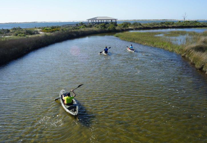 Three kayakers paddle around coastal marsh with view of Pavilion in the background. 