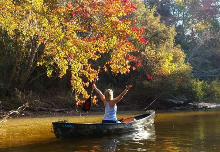 Woman in canoe throws arms up to embrace her surroundings. Fall foliage is a mix of vibrant yellows and oranges. 