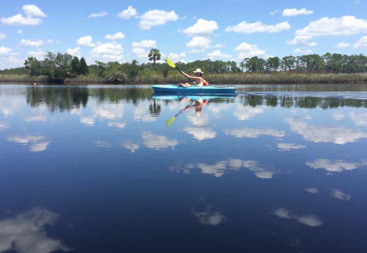 Woman paddles through water so calm it is reflecting the clouds and sky. 