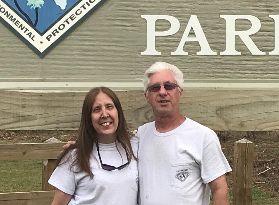 Volunteer duo Gene and Annelise Carlson smile proudly in front of the park entrance sign. 