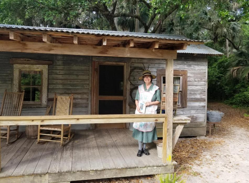 a woman in period dress stands on the porch of a wooden cabin