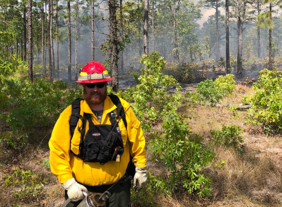 A ranger in firefighting gear holds a drip torch while woods burn in the background.