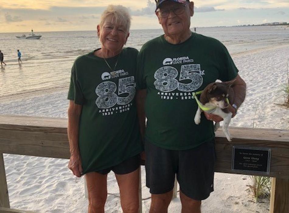 Two volunteers wearing Florida State Parks 85th Anniversary t-shirts, smiling at the camera and holding a little dog, with the beach in the background.