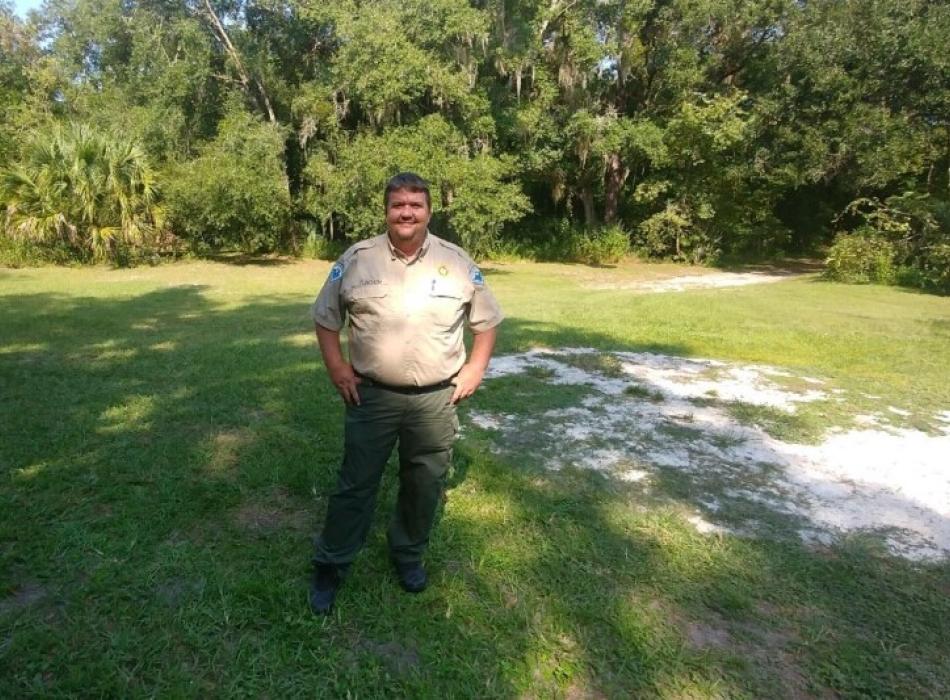 A man in uniform stands in front of a row of trees. 