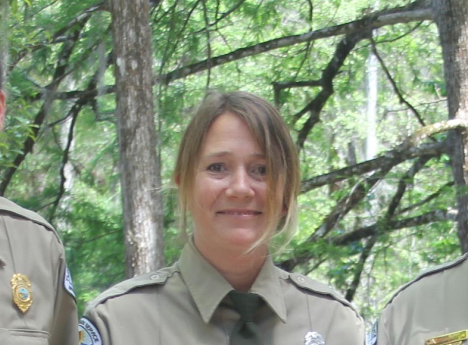 a blonde woman stands in front of trees in a park service uniform
