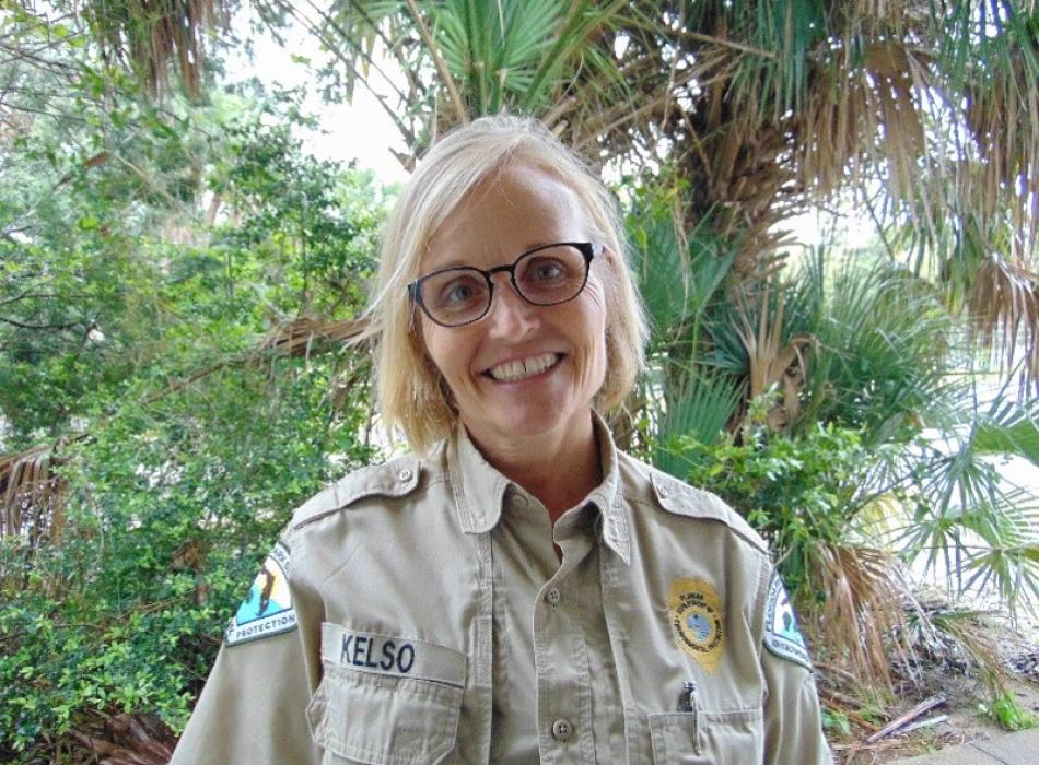 a woman in park service uniform and glasses smiles at the camera in front of a palm tree.